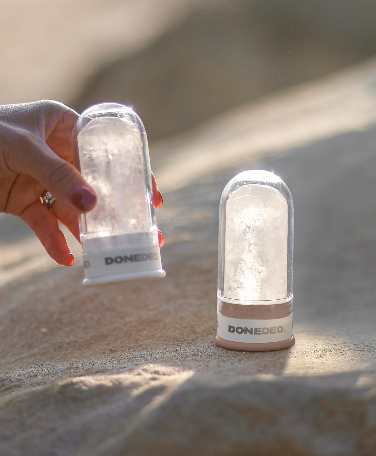 Limited edition DONEDEO natural deodorant sticks on sandy surface, with one held by hand and one standing upright in warm natural light